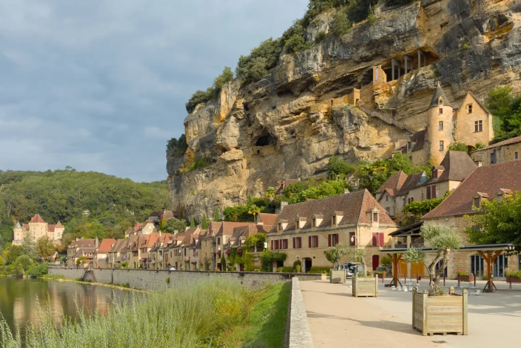 La Roque-Gageac — Les maisons aux toits bruns alignées au pied de la grande falaise, vues depuis la rive.