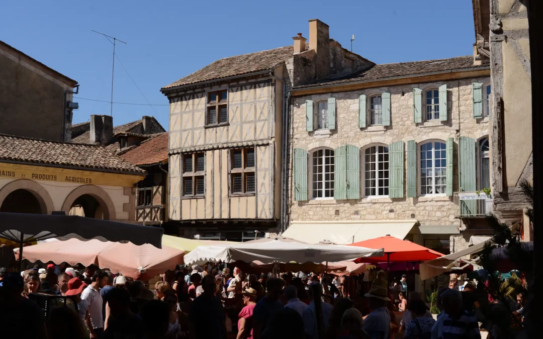 Le marché du dimanche sur la place de l'église d'Issigeac, parasols et maisons à pans de bois sous un ciel bleu